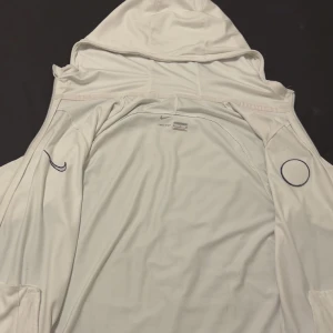 Vit PSG hoodie från Nike - Säljer en vit hoodie från Nike med PSG-logga på bröstet och blå Nike-swoosh. Jackan har dragkedja, huva med dragsko och är tillverkad i ett mjukt, sportigt material. Slim fit-modell som passar perfekt till fotboll eller streetwear.