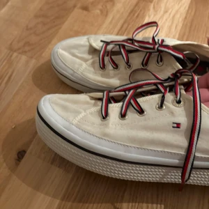 Vita sneakers från Tommy Hilfiger - Säljer ett par vita sneakers från Tommy Hilfiger med klassiska Hilfiger-detaljer på sidan och på innersulan. Skorna har snörning med röd, vit och blå randiga skosnören och en platt sula.