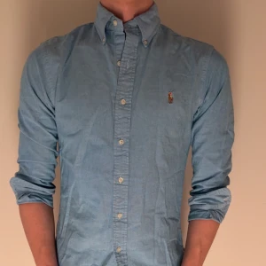 Ljusblå skjorta från Ralph Lauren - Klassisk ljusblå skjorta från Ralph Lauren med button-down krage och broderad logga på bröstet. Skjortan har långa ärmar och knäppning framtill. Perfekt för en stilren och avslappnad look.