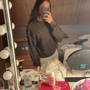 Brun stickad tröja med rund hals - Mysig brun stickad tröja med rund hals och diskreta håldetaljer längs axlar och bröst. Tröjan har lång ärm och en avslappnad passform, perfekt för kyligare dagar.