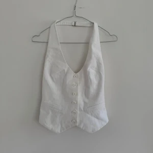 Vit väst med djup v-ringning från mango - Säljer en stilren vit väst från Mango med djup v-ringning och knappar framtill. Västen har två dekorativa fickor och är figursydd för en snygg passform. Öppen rygg!