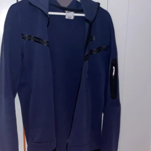 Mörkblå zip-hoodie från Nike - Mörkblå hoodie från Nike med dragkedja och huva. Hoodien har svarta detaljer på bröstet och en ficka med dragkedja på ärmen. Perfekt för en sportig stil och enkel att matcha.