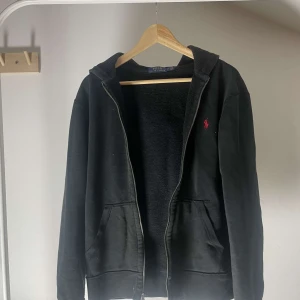 Svart hoodie med dragkedja från Polo Ralph Lauren - Svart hoodie från Polo Ralph Lauren med dragkedja framtill och huva. Tröjan har två fickor och en röd broderad logga på bröstet. Mjuk insida och klassisk passform, perfekt för en avslappnad stil.