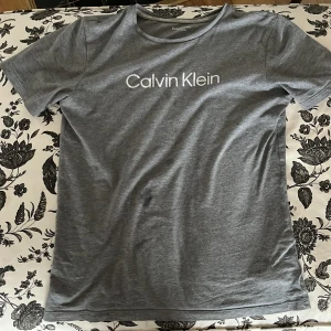 Grå t-shirt från Calvin Klein - Säljer en stilren grå t-shirt från Calvin Klein med vit logga tryckt på bröstet. T-shirten har rund halsringning och är kortärmad. Perfekt för en enkel och snygg look. Den har kanske 1 fläck men inte alls stor den stora fläcken e bara vatten!