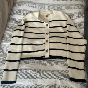 Randig croppad kofta från Zara - Säljer en vit och svart randig croppad kofta från Zara. Tröjan har långa ärmar, ribbad struktur och snygga knappar framtill. Perfekt för dig som gillar en stilren och trendig look.