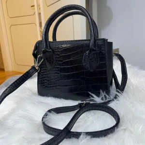 Svart axelväska med krokodilpräglat mönster - Snygg svart axelväska i syntetmaterial med krokodilpräglat mönster. Väskan har både handtag och justerbar axelrem, perfekt för att bära på olika sätt. Stilren och trendig design som passar till många outfits.