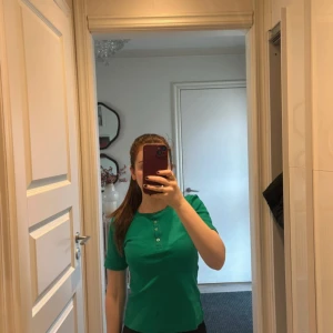 Grön ribbad topp med knappar - Säljer en grön ribbad topp från Cubus i storlek XS. Toppen har korta ärmar, rund halsringning och fyra knappar framtill.