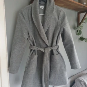 Grå kappa från Bubbleroom - Snygg grå kappa från Bubbleroom med krage och bälte i midjan. Kappan har fickor framtill och stängs med knappar. Inga skador eller defekter förekommer, storlek 36🩷
