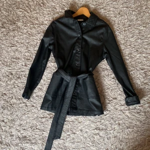 Svart skinnjacka med bälte - Snygg svart skinnjacka med knappar framtill och tillhörande bälte i midjan. Jackan har lång ärm och klassisk krage, perfekt för en stilren look. Passformen är normal. Använd bara en gång💖