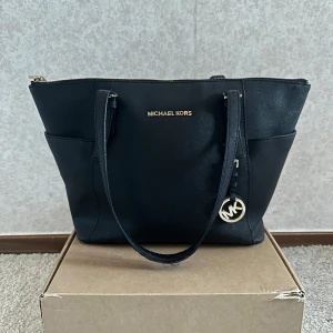 Svart axelväska från Michael Kors - Stilren svart axelväska från Michael Kors med guldfärgade detaljer och logotyp framtill. Väskan har dragkedja upptill, två långa axelband och en rund MK-hänge i metall. Lite slitet på axelbanden!