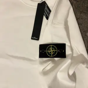 Säljer en stilren vit sweatshirt från Stone Island med klassisk rund hals och den ikoniska logotypen på vänster ärm. Tröjan har långa ärmar och ribbade muddar vid ärmslut och nederkant. Perfekt för en clean och avslappnad look.