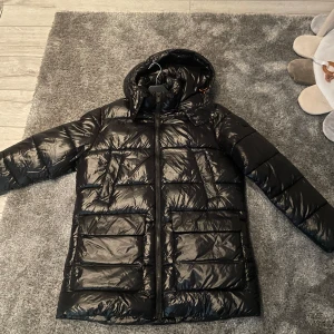Svart dunjacka med huva från Save The Duck - Säljer en svart pufferjacka från Save The Duck med huva och stora fickor framtill. Jackan har dragkedja och är tillverkad i syntetmaterial, helt fri från animaliska produkter. Perfekt för kyliga dagar. Nytt skick!! Pris kan diskuteras! 