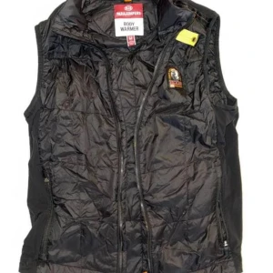 Svart dunväst från Parajumpers - Säljer en svart dunväst från Parajumpers med dragkedja framtill och klassisk logga på bröstet. Västen har hög krage och en gul detalj vid axeln. Perfekt för lager-på-lager och håller dig varm under kyliga dagar.