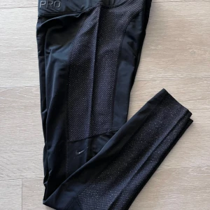 Svarta träningstights från Nike Pro - Svarta tights från Nike Pro med meshpaneler längs benen för extra ventilation. Tight passform och elastisk midja. Perfekta för gymmet eller löpning. Diskret Nike-logga på benet och PRO-tryck i midjan.