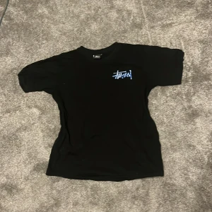 Svart t-shirt från Stüssy med blått tryck - Svart t-shirt från Stüssy i 100% bomull med blått och vitt Stüssy-tryck på bröstet och större tryck på ryggen. Klassisk passform och korta ärmar. Perfekt för dig som gillar streetwear och vill sticka ut med en snygg logga.