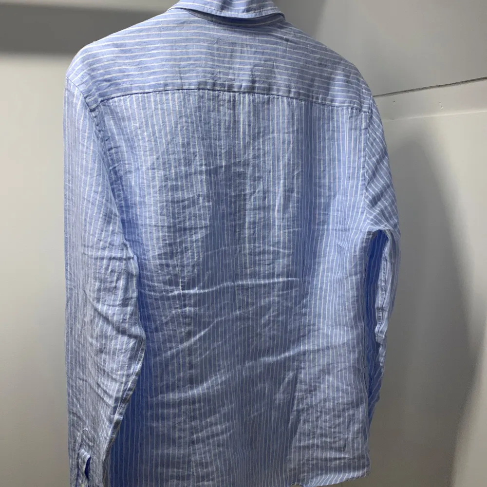 Snygg ljusblå skjorta från Bläck med vita ränder och klassisk krage. Skjortan har lång ärm, knappar framtill och är i en skräddad passform (tailored fit). Perfekt för dig som gillar stilrena och fräscha plagg.. Paidat.