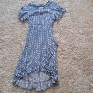 Blommig blå omlottklänning från SRN HLT - Säljer en blå långklänning från SRN HLT med vitt blommönster. Klänningen har korta ärmar, volangdetaljer och omlott med knytband i midjan. Perfekt för sommarens tillfällen!