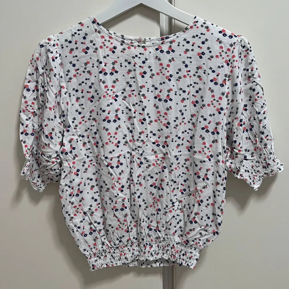 Söt vit blus med små blommor i rosa, blått och grått. Blusen har korta puffärmar med resår och resår i midjan för en feminin siluett. Perfekt för vår och sommar, lätt och luftig i bomull.. Yläosat.