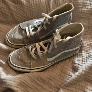 Gråa höga sneakers från Vans - Säljer ett par gråa höga sneakers från Vans med vit sula och vit sidodetalj. Skorna har snörning och är tillverkade i mocka och textil. Klassisk Vans-logga baktill på sulan. Sparsamt använd 