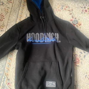 Svart hoodie från Hoodrich med blå detaljer - Svart hoodie från Hoodrich med stor logga i vitt och blått framtill. Tröjan har huva med blått foder, dragsko och en stor magficka. Perfekt för en avslappnad stil.