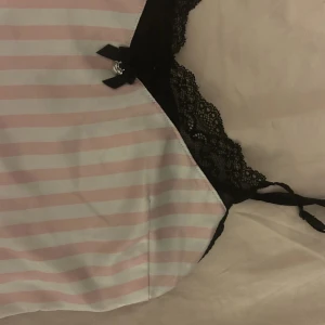 Randig silkes pyjamas med spetsdetaljer - Säljer en söt pyjamas set med rosa och vita ränder samt svarta spetsdetaljer upptill och under bysten. Toppen har tunna axelband och en liten rosett framtill. Perfekt för en trendig och feminin look.