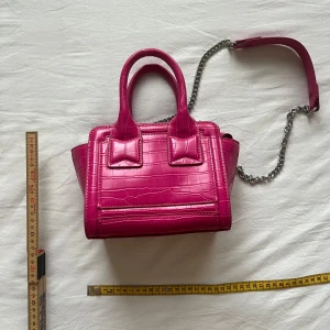 Rosa handväska med kedjerem - Säljer en snygg  rosa handväska med krokodilpräglat mönster och silverfärgad kedjerem. Väskan har två handtag och stängs med dragkedja. Perfekt accessoar för att lyfta din outfit!