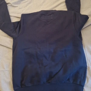 Mörkblå sweatshirt från Henri Lloyd - Säljer en mörkblå sweatshirt från Henri Lloyd med rund halsringning och diskret logga tryckt på bröstet. Tröjan har långa ärmar och ribbade muddar vid ärmslut och nederkant. Perfekt för en avslappnad stil.