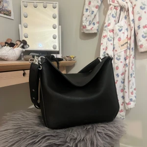 Svart axelväska  - Säljer en stilren svart axelväska i skinn med bred axelrem och silverfärgade detaljer. ☺️ Väskan har en enkel, modern design och är rymlig med praktisk öppning upptill. Perfekt för dig som vill ha en klassisk accessoar till vardagen. Finns två olika band som båda går att justera, väskan är i gott skick skriv för fler bilder. Från Rizzo ca 1000 kr minns inte exakt, märket har gått i konkurs.