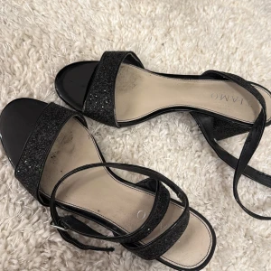 Svarta glittriga sandaler med klack från Tiamo - Säljer ett par svarta sandaler från Tiamo med glittriga remmar och bred klack. Skorna har öppen tå och justerbart spänne runt vristen som funkar bra. Perfekta för bal eller en fest! De är använda fåtal gånger men har fortfarande kvar sin bling!
