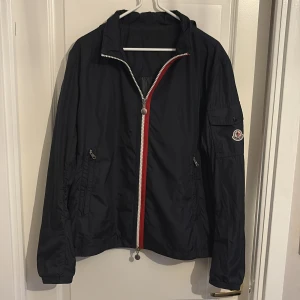 Moncler vårjacka - Säljer en mörkblå vindjacka från Moncler med huva som även går fälla in.  Perfekt för blåsiga dagar nu till vår tidig sommar. Storlek Xl men sitter som M eftersom relativt kort. Hör av vid funderingar 