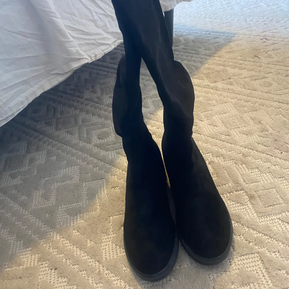Säljer ett par svarta höga stövlar i mjuk mocka med platt sula. Skorna har rund tå och är perfekta för att ge en stilren look till din outfit. Passar bra till både byxor och kjol.. Kengät.