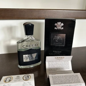Creed Aventus Eau de Parfum 100ml - Exklusiv herrparfym från Creed, Aventus, i en elegant glasflaska med svart etikett och silverdetaljer. Flaskan rymmer 100 ml och kommer med originalkartong och informationskort. Doften är känd för sin fräscha och maskulina karaktär.