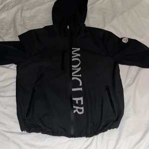 Windbreaker från Moncler - Säljer en svart vindjacka från Moncler. Den har  en bröstficka med dragkedja. Fodret är slätt och insidan har Moncler-etikett. Perfekt för blåsiga dagar. Den är lite lös där man spänner!!