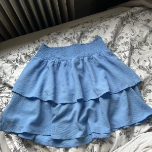 Blå volangkjol från ONLY - Supersöt blå kjol från ONLY med dubbla volanger och smock i midjan. Kjolen har en luftig känsla, perfekt för varma dagar. Enkel att matcha med olika toppar för en fräsch look.