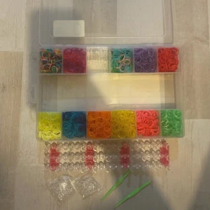 DIY elastikarmbandskit från Tiger - Säljer ett komplett DIY-kit för att göra egna elastiska armband. Kitet innehåller färgglada gummiband i flera färger, två gröna krokar, plastclips och en armbandsväv. Perfekt för att skapa personliga smycken!
