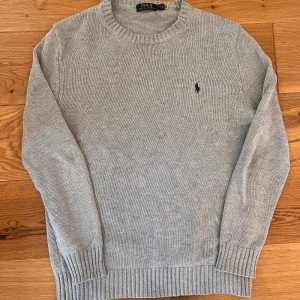 Grå stickad tröja från Polo Ralph Lauren - Säljer en klassisk grå stickad tröja från Polo Ralph Lauren med rund halsringning och broderad logga på bröstet. Tröjan har långa ärmar och ribbade muddar vid ärmslut och nederkant. Perfekt för lager-på-lager under kyliga dagar. Obs inte orginal