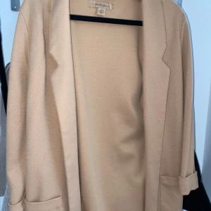 Beige kavaj från H&M - Säljer en stilren beige kavaj från new yorker med klassisk krage och slag. Kavajen har långa ärmar med uppvik och två fickor framtill. Perfekt för en snygg och enkel look.