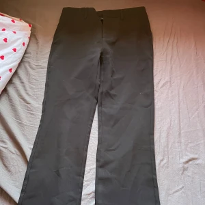 Svarta kostymbyxor med raka ben - Säljer ett par klassiska svarta kostymbyxor med raka ben och diskreta fickor bak. Byxorna har hällor för bälte och stängs med dragkedja och knapp framtill. Perfekta till en stilren look.