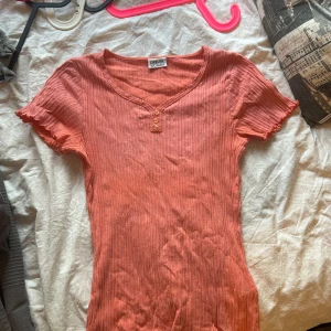 Koralrosa ribbad topp från Casual Woman - Säljer en korallrosa ribbad topp från Casual Woman med v-ringning och tre små knappar framtill. Toppen har korta ärmar med vågig kant och är tillverkad i 100% bomull. Perfekt för en avslappnad stil.