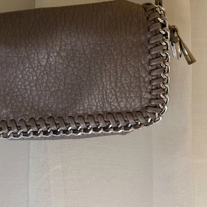 Grå axelväska med kedjedetaljer - Säljer en snygg grå axelväska i skinnimitation med silverfärgad kedja som axelrem och runt kanterna. Väskan har magnetlås och ett stort fack inuti. Perfekt för dig som vill ha en stilren accessoar med lite edge. Jag säljer för 89 kr + frakt!