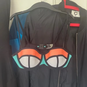 Färgglad bikiniöverdel från Triangl - Säljer en bikiniöverdel från Triangl i färgerna orange, turkos och svart med grafiskt mönster. Överdelen har bygel och justerbara axelband samt spänne i ryggen. Perfekt för sommaren!