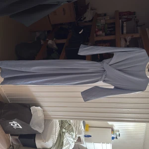 Ljusblå långklänning med knytband - Säljer en elegant ljusblå långklänning med långa ärmar och knytband i midjan. Klänningen har en omlott-look upptill och är perfekt för dig som vill ha en stilren och enkel look.