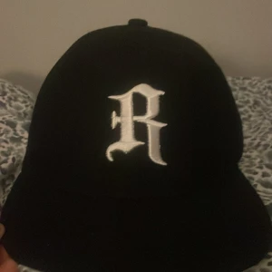 Svart keps med broderad R och Golden Age-märke - Svart keps med vit broderad bokstav 'R' framtill och färgglatt 'Golden Age' märke på sidan. Justerbar snapback baktill för flexibel passform. Perfekt accessoar för en cool streetwear-look.