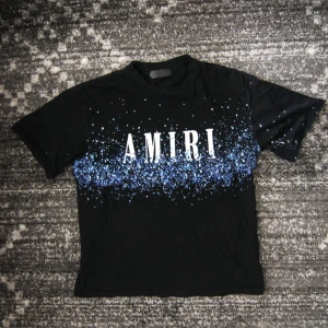 Svart Medium t-shirt från Amiri med blått stänkmönster - Säljer en svart t-shirt från Amiri med stort vitt tryck framtill och blått stänkmönster över bröstet och ryggen. T-shirten har rund hals och korta ärmar. Perfekt för dig som gillar streetwear och vill sticka ut.