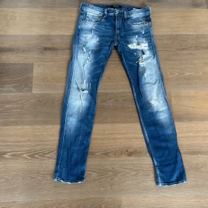 Blå Replay Anbass jeans med slitningar - Säljer ett par blå Replay Anbass jeans med tydliga slitningar och ljusa partier. Jeansen har klassisk femficksdesign, dragkedja och knapp i midjan. Passformen är smal och materialet är denim med stretch. Priset är inte hugget i sten. Hör av er för frågor och prisförslag.