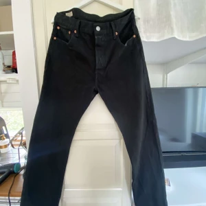 Svarta Levi's 501 jeans - Klassiska svarta Levi's 501 jeans med raka ben och knappgylf. Byxorna har fem fickor och den ikoniska läderpatchen bak i midjan. Perfekta för dig som gillar tidlös stil och vill ha ett par jeans som funkar till allt.