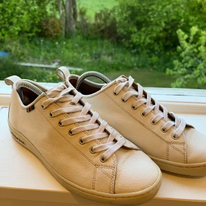 Vita sneakers från PS Paul Smith - Säljer ett par stilrena vita sneakers från PS Paul Smith. Skorna har rund tå, platt sula och klassisk snörning. Tillverkade i skinn med diskret logga på sidan och små färgdetaljer vid snörningen. Perfekta för en clean och enkel look. Lite smutsiga snören som går att tvätta bort samt märket på höger skol har försvunnit lite