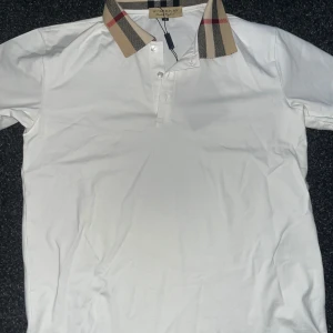 Vit pikétröja från Burberry  - Säljer en vit pikétröja från Burberry med den ikoniska rutiga kragen i beige, svart och rött. Tröjan har korta ärmar och knäppning framtill med tre knappar. Perfekt för en stilren och klassisk look. Helt oanvänd 