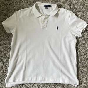 Vit pikétröja från Polo Ralph Lauren - Klassisk vit pikétröja från Polo Ralph Lauren med blå broderad logga på bröstet. Tröjan har korta ärmar, krage och knappar framtill. Perfekt för en stilren och tidlös look. 68 cm från axel och ner. 