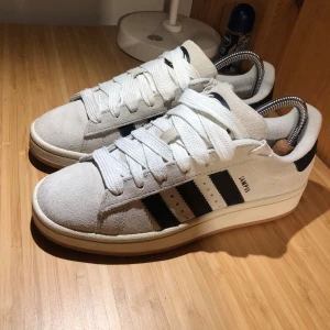 Adidas Campus vita sneakers med svarta ränder - Säljer ett par klassiska Adidas Campus sneakers i vitt med svarta ränder på sidorna och beige sula. Skorna har breda vita skosnören och är i en stilren design som passar till det mesta. Storlek 39 1/3. Helt nya, endast använda enstaka gång